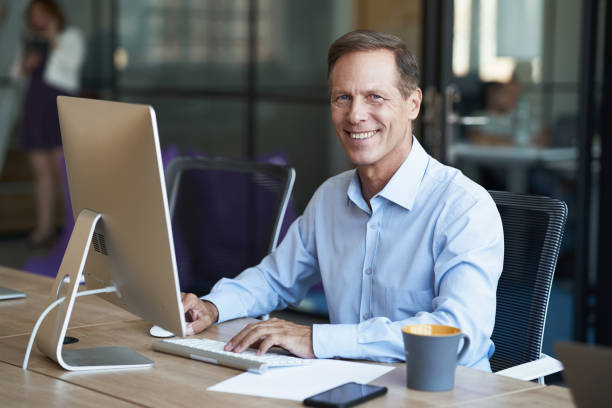 istock Cheerful mature businessman sitting at desk in the modern office and working on computer, smiling at camera 1287095687