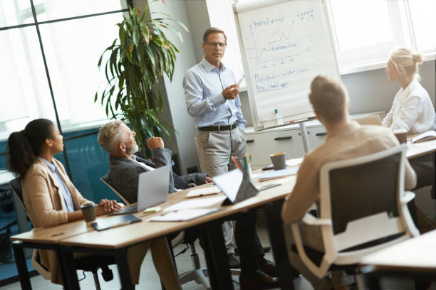 istock Confident mature businessman standing against flipchart and explaining new business strategy to colleagues while having a meeting in the modern office 1287096404