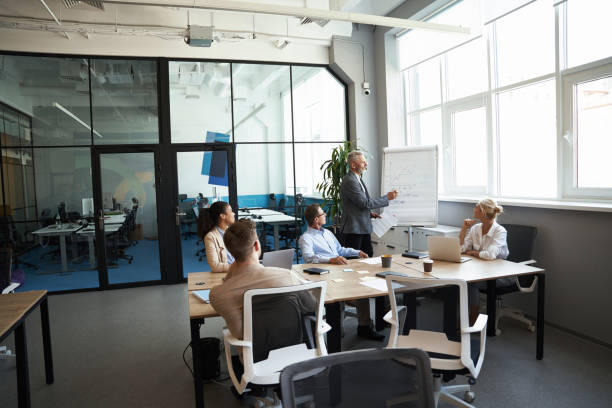istock Startup team having a meeting. Confident mature businessman writing something on flipchart while sitting with multiracial colleagues in the modern office. 1287096434