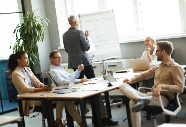 istock Confident mature businessman writing something on flipchart while his colleagues discussing something, having a meeting in the modern office 1287096454