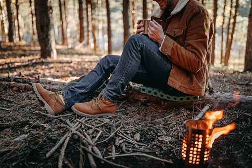 Young man sitting by fire and enjoying hot tea while camping in forest