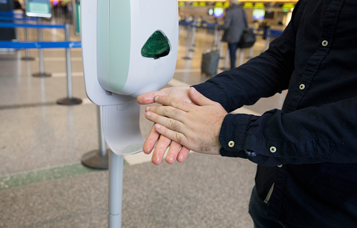 Man at the airport washes his hands using hand sanitizer dispenser in the airport to combat flu, cold and coronavirus.