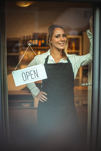 Portrait of a smiling Caucasian woman with black apron, standing behind glass door