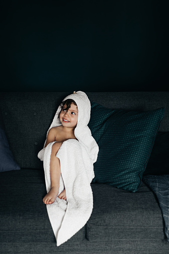 A cute little girl sitting in the living room wrapped in a big white towel with a hood after taking a bath.