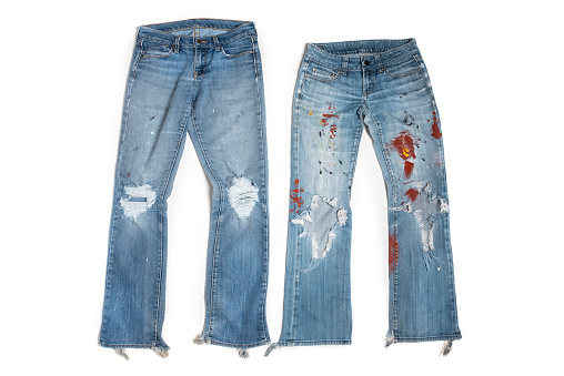 Horizontal photograph of two pair of worn out blue jeans. They are positioned on a white background. Both jeans have large holes on the knees and the bottom hem of the legs are torn. One pair of jeans has splatters of paint on the legs.