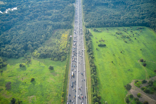 top-down luftaufnahme von drohne der langen geraden mehrspurigen autobahn mit stau auf der straße und zwischen grüner natur. sonnenlicht, wald und grüne felder rund um die straße. kamera schaut schräg nach unten. - driving industry land vehicle multiple lane highway stock-fotos und bilder