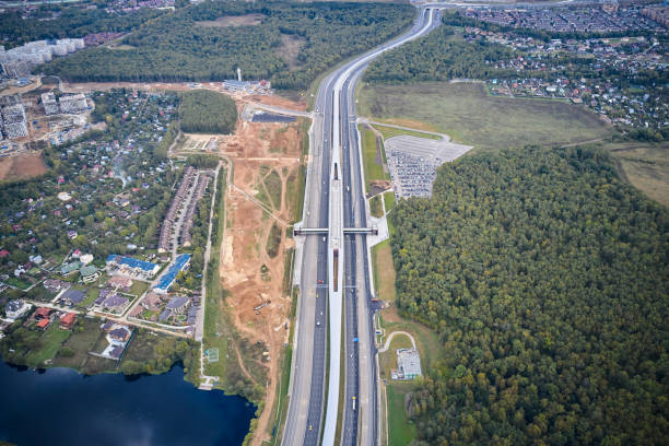 luftaufnahme der langen geraden und kurve mehrspurige autobahn mit u-bahn-linie in der mitte. waldgebiete, see- und vorortwohnviertel. hohe höhe und weit weg geschossen. autos sind unterwegs. kamera schaut schräg nach unten - driving industry land vehicle multiple lane highway stock-fotos und bilder