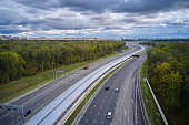 High angle view of the long turning multiple lane highway with the metro line in the center. The way to city between the green forest all around. A few cars are crossing the highway at other sides. Camera looks down at an angle. Cityscape on background.