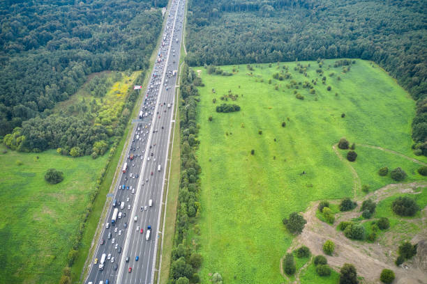 top down aerial view from drone of the long straight multiple lane highway with traffic jam on the road between green meadows and forest. vertical road on the left part of photography. camera looks down at an angle. - multiple lane highway highway car field imagens e fotografias de stock