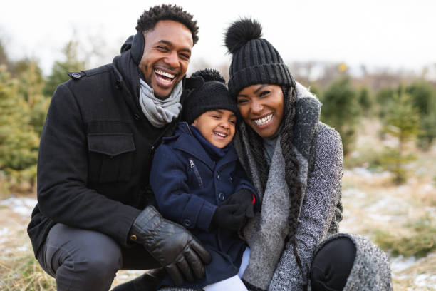 A portrait of a young family at a Christmas tree farm A young attractive African American family pose for a photo at a tree farm. They are all wearing warm winter clothes and smiling directly at the camera. The young girl is of elementary school age. vacations photos stock pictures, royalty-free photos & images