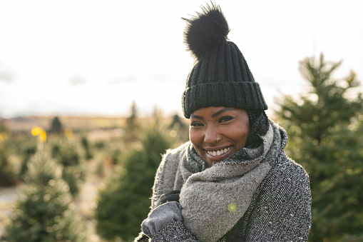 A portrait of a beautiful women of African American descent is smiling. She is wearing warm winter clothes and is a tree farm.