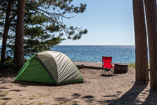 A green tent is located on the beach during summer. The camping spot offers a beautiful view of Lake Superior. It is located in Lake Superior Provincial Park in Ontario, Canada. A red chair is next to the fireplace.
