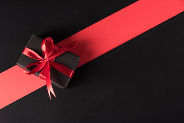 Black Friday sale shopping and Boxing Day concept, Gift box wrapped black paper and red bow ribbon present, studio shot isolated on red and dark background