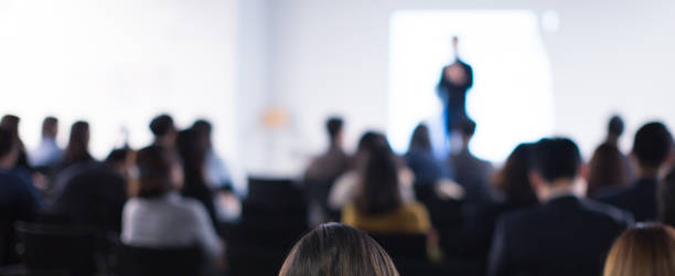 Speaker on the stage with rear view of audience in the conference hall or seminar meeting, business and education concept. Speaker giving a talk at business meeting. Seminar presentation photo. stock photo