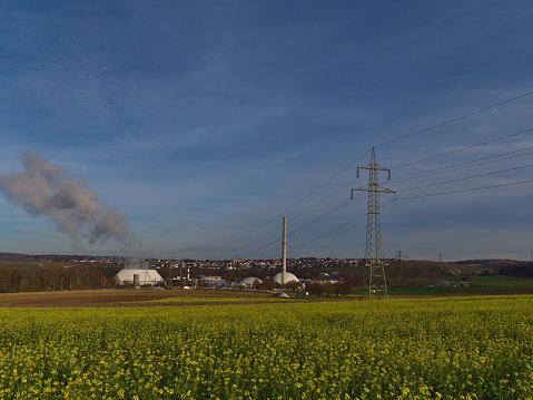 Nuclear power plant in Neckarwestheim, Germany, located at the bank of Neckar river, with power line, pylon and a blooming agricultural field with yellow rapeseed flowers in front in fall season.