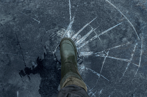 Fisherman foot on broken cracked thin ice at lake. Dangerous winter fishing