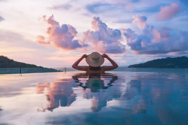 Summer travel vacation concept, Happy traveler asian woman with hat and bikini relax in luxury infinity pool hotel resort with sea beach at sunset in Phuket, Thailand