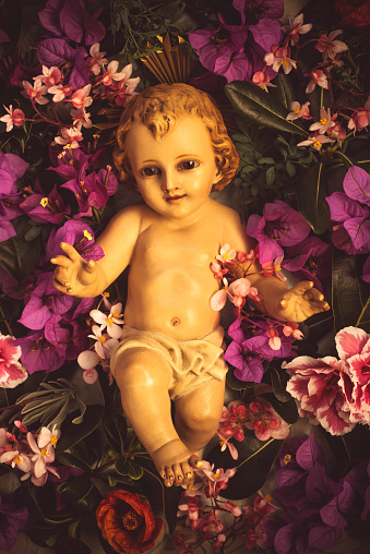 Old figurine of the Christmas Child Jesus surrounded by flowers of different varieties, retro style, vertical image.