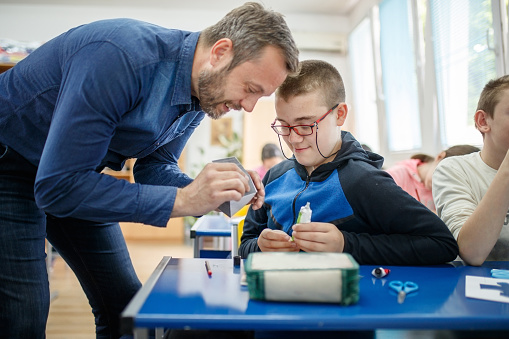 Male special education teacher working closely with a intellectual disabiled elementary age boy, he is showing him to glue cut paper following pattern while boy with eyeglasses is watching