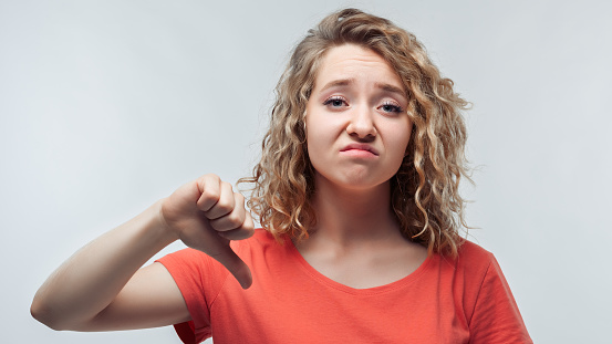 Pretty blonde girl with curly hair in casual t shirt showing thumb down. Unlike, dislike, failure gestures concept. Studio shot, white background. Human emotions, facial expression concept