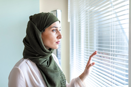 Modern and Young Muslim Woman is Thoughtfully Standing Near the Window in the Domestic Room. Arabian Ethnicity woman is covered with a Traditional Scarf and Looking Through the Window Glass.