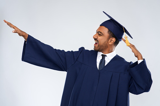 education, graduation and success concept - happy smiling indian male graduate student in mortar board and bachelor gown celebrating success over grey background