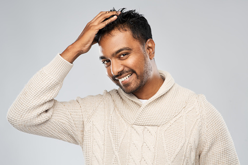 emotion, expression and people concept - smiling indian man in knitted woollen sweater touching his hair over gray background