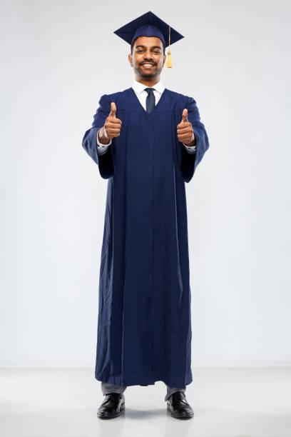 education, graduation and people concept - happy smiling indian male graduate student in mortar board and bachelor gown  showing thumbs up over grey background