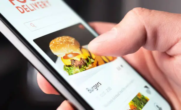 Photo of Food delivery app order with phone. Online mobile service for take away burger and pizza. Hungry man reading restaurant menu, website and reviews with smartphone.