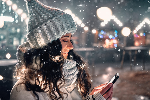 Pretty girl wearing a knitted scarf and hat holding a smartphone. Merry Christmas and Happy New Year to your loved ones and family. Festive night city lights and garlands background.