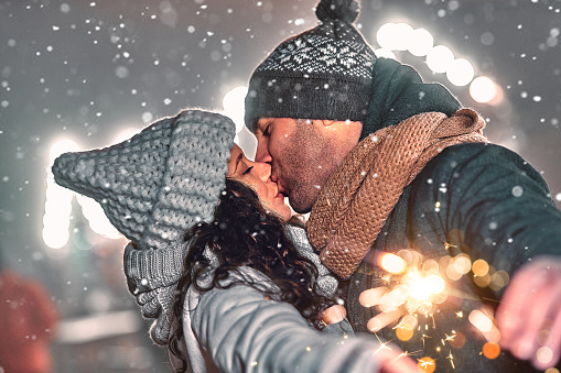 A loving couple dressed in knitted hats and scarves celebrate the New Year against the backdrop of the night city, hold sparklers, it snows and there is a magical atmosphere