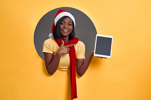 A young African woman in a santa claus hat and red scarf smiles and points to a tablet with empty space. Discounts, gifts, benefit concept.