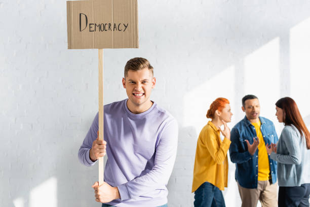 istock angry man grimacing while holding placard with democracy lettering near multicultural people talking on blurred background 1287206169