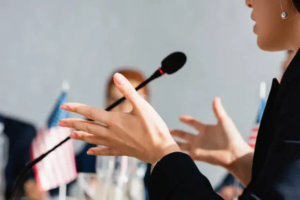 Photo of Cropped view of female politician gesturing, while speaking in microphone with blurred woman on background