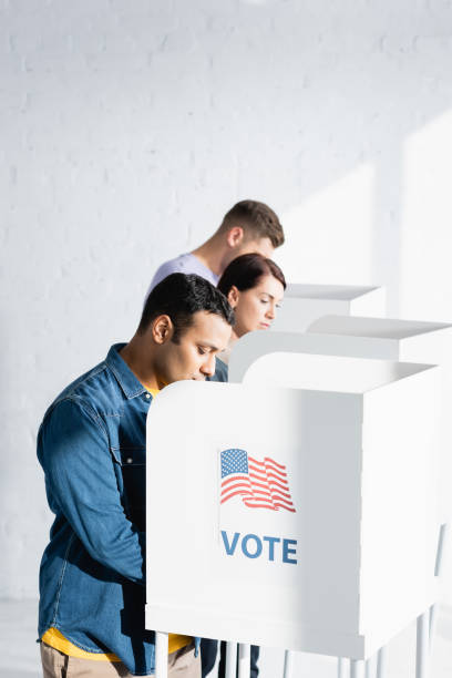 multicultural voters in polling cabins with american flag and vote inscription on blurred background - jovens a votar imagens e fotografias de stock