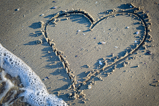 Stock photo showing close-up view of a pile of seashells with a starfish surrounding a sandstone heart standing up in the sand on a sunny, golden beach with sea at low tide in the background. Romantic holiday and honeymoon concept.