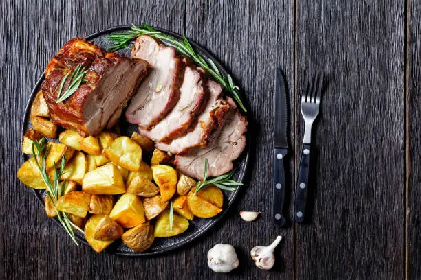 Sliced roasted pork loin served with baked potato wedges, rosemary  on a plate with garlic steak cutlery on a dark wooden background, top view, close up