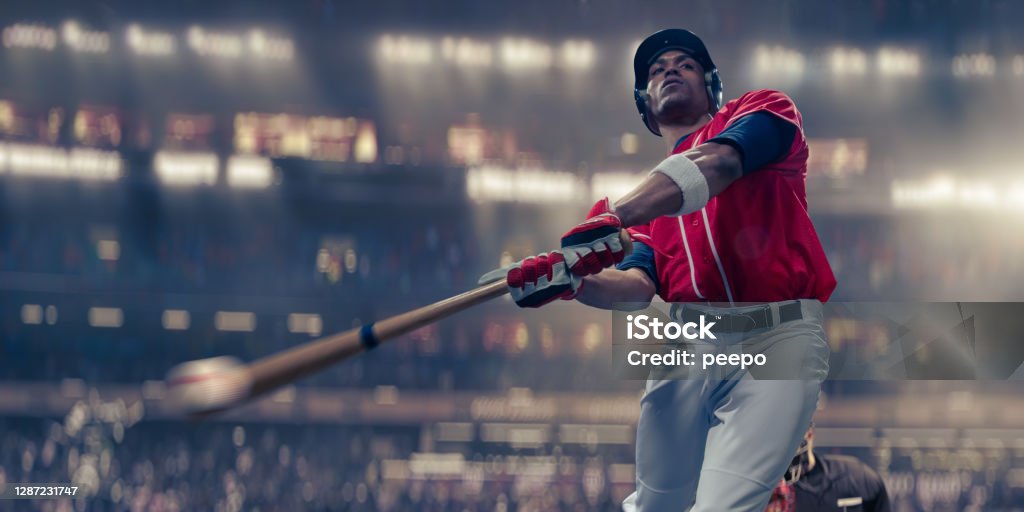Professional Baseball Batter Hitting Baseball In Mid-Swing Close Up A close up of a professional baseball batter in mid swing, having just struck the baseball. The athlete wears a red baseball jersey and white trousers, gloves and safety helmet. The baseball player is playing in a generic floodlit stadium. With selective focus and motion blur to bat and ball. Baseball - Sport Stock Photo