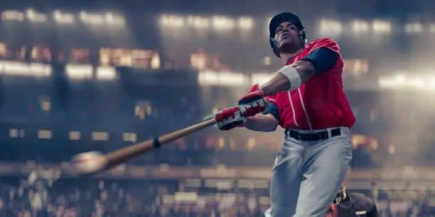A close up of a professional baseball batter in mid swing, having just struck the baseball. The athlete wears a red baseball jersey and white trousers, gloves and safety helmet. The baseball player is playing in a generic floodlit stadium. With selective focus and motion blur to bat and ball.