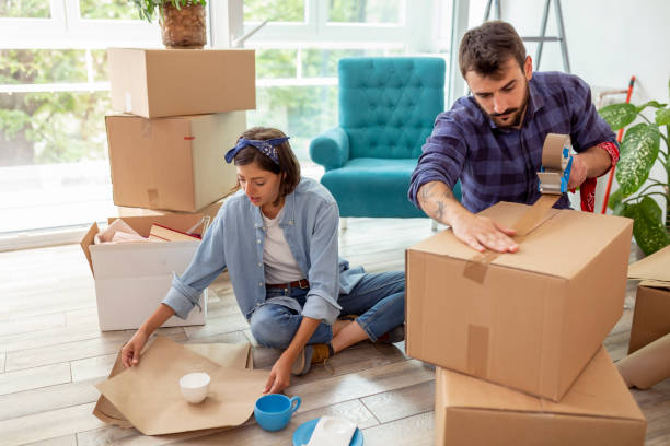 Couple moving house, packing things Couple in love packing things into cardboard boxes, getting ready for relocation - man taping boxes using packing machine while woman is wrapping fragile things into paper packing stock pictures, royalty-free photos & images