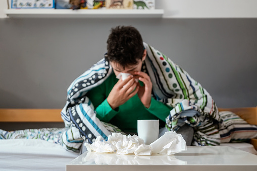 Sick teen boy sitting in bed drinking tea and blowing nose