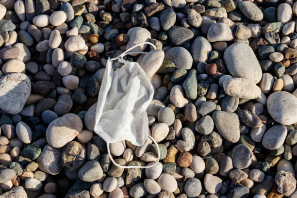 sujo usou máscara médica descartável em uma praia de pedras à beira-mar. resíduos durante o covid-19. poluição devido à pandemia coronavírus - 5428 - fotografias e filmes do acervo