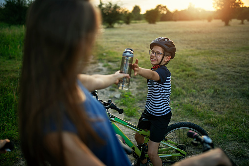 Happy little boy riding bicycle on a hot summer day. The sister is giving him reusable water bottle.\nNikon D850