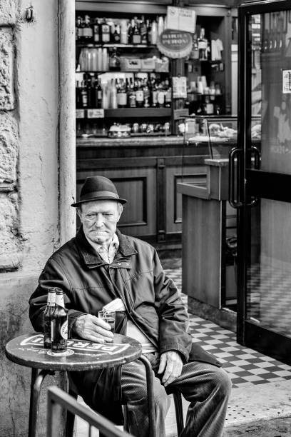 An elderly man sitting outside a bar in the ancient Trastevere district in the heart of Rome Rome, Italy, October 28 -- A lonely old man drink a beer sitting outside a bar in the ancient Trastevere quarter, the most loved and visited roman district by tourists and residents of the eternal city, with a lot of restaurants and tavern of Roman and Italian cuisine. Image in High Definition format. italian language stock pictures, royalty-free photos & images