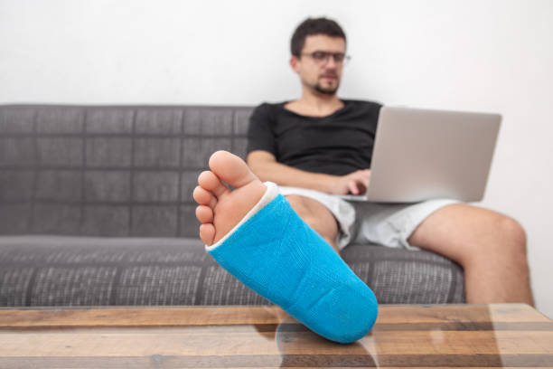 istock Man with glasses with broken leg in cast working on a laptop on couch at home. 1287249903