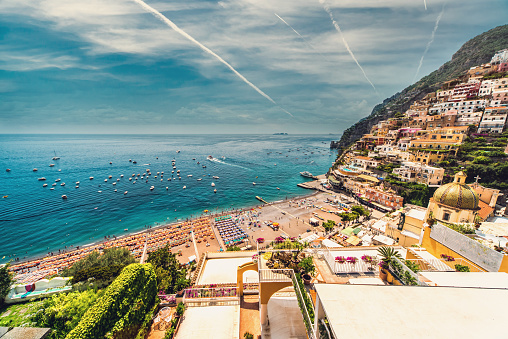 Amazing Amalfi coast with beach with parasols, hillside architecture, bright sky, view from above, jets leave white trail in cloudy sky. European resort, famous touristic place. Europe Positano, Italy