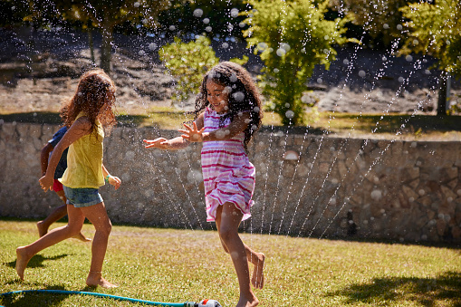 Small african-american girl jumping over sprinkler on garden with friends