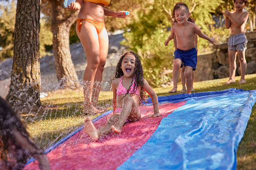 Group of multi-ethnic children sliding on water slide with mother on garden
