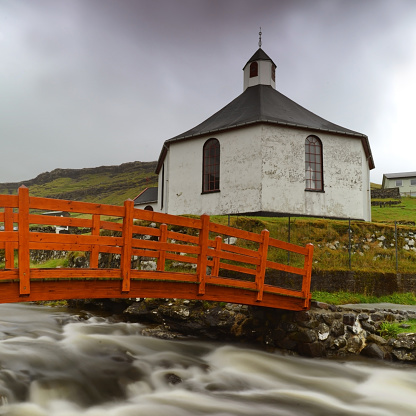 Fisheye lowangle shot of Haldorsvíkar Church with orange bridge and small river taken with long exposure on a cloudy rainy day in Haldorsvik, Faroe Islands.