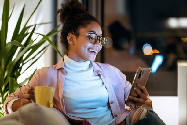 joven empresaria de felicidad que usa su teléfono inteligente mientras bebe una taza de café sentado en el sofá en la oficina. - mobile marketing fotografías e imágenes de stock